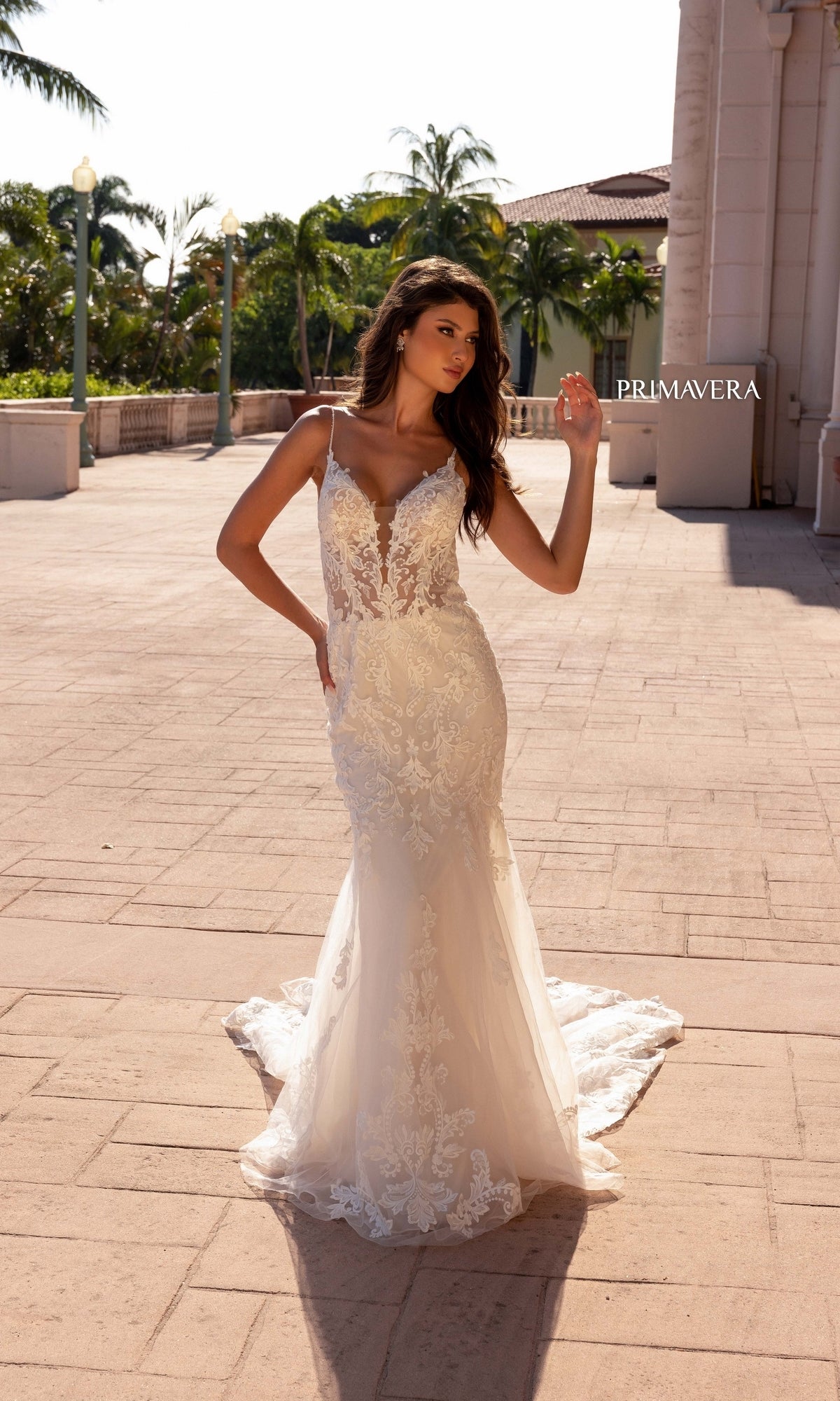 A woman poses outdoors on a sunlit patio beside palm trees and a building, wearing the Long Wedding Dress 11137 by Primavera—an ivory, fitted gown with lace embroidery and thin straps.